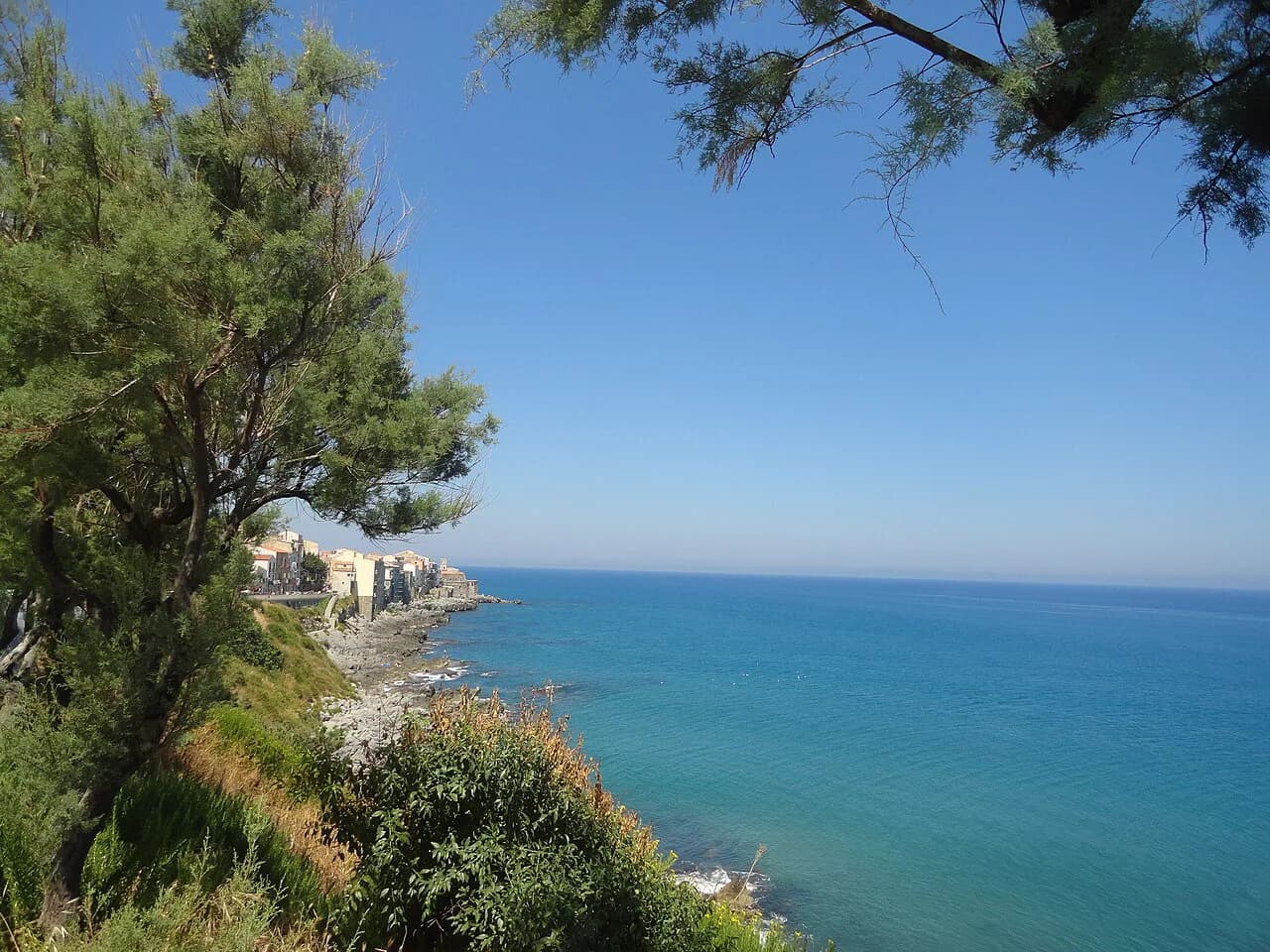 Pine trees and green shoreline above a turquoise bay, with Cefalù's old town in the distance