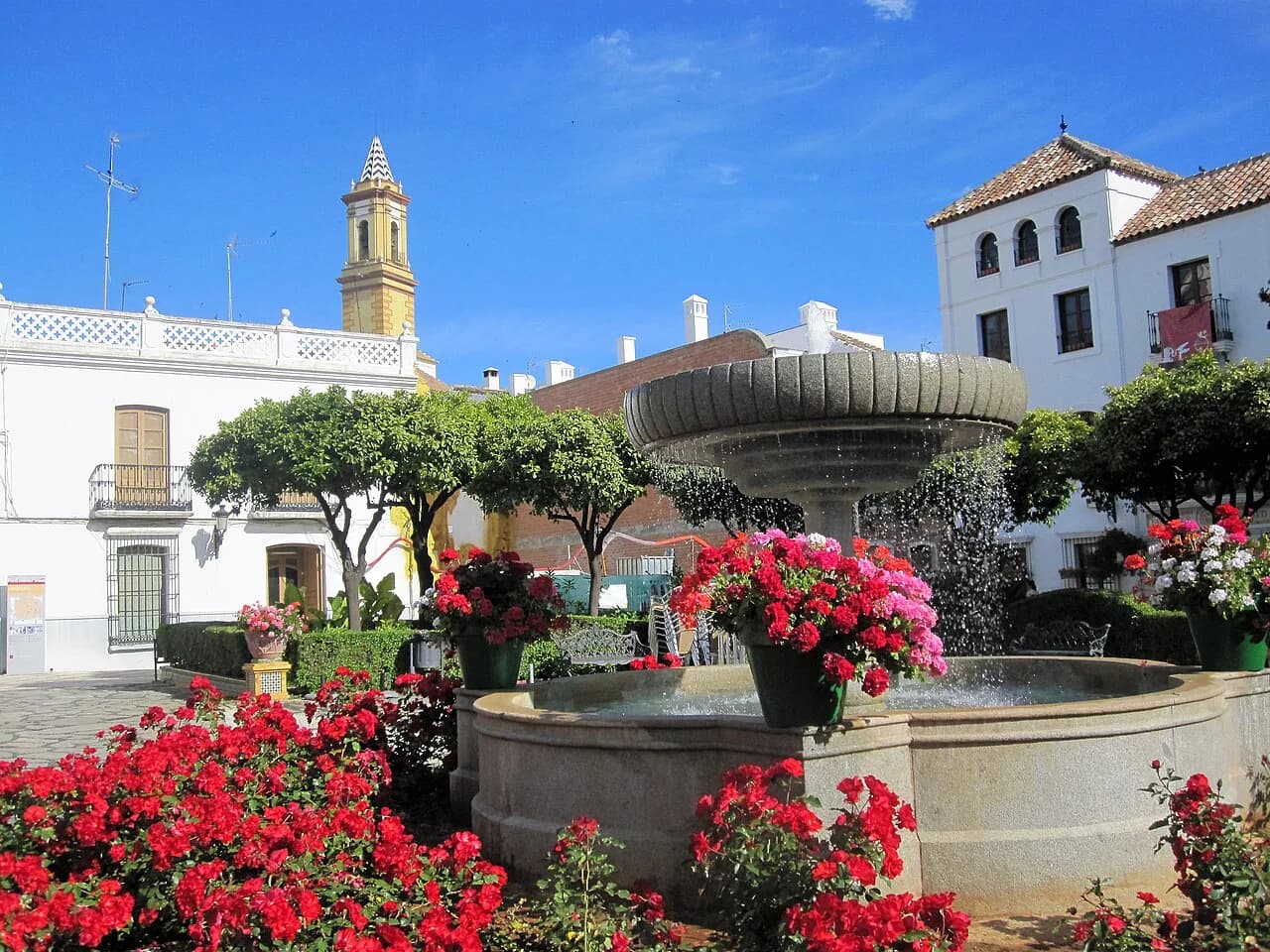 Fountain and red flowers in Plaza de las Flores with a bell tower behind, Estepona
