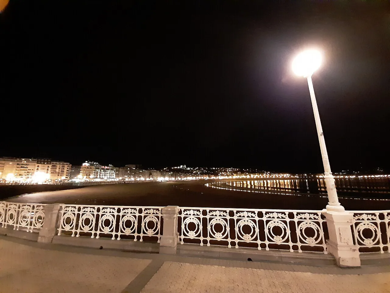 The white wrought-iron railing and lamp of the La Concha promenade at night, with the lit bay beyond, San Sebastián