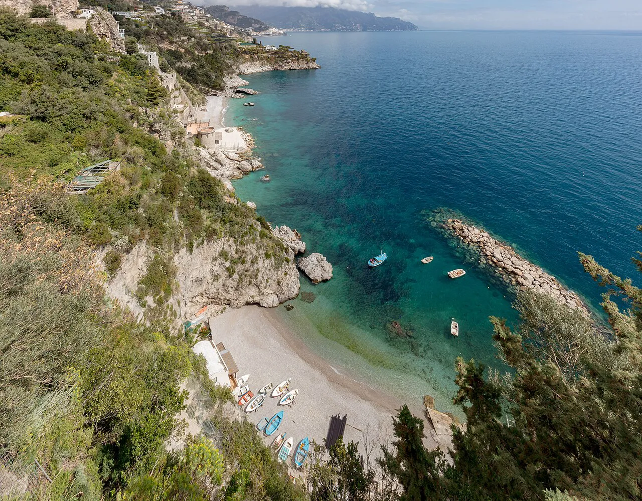 Small pebble cove with turquoise water and moored boats below green cliffs at Conca dei Marini