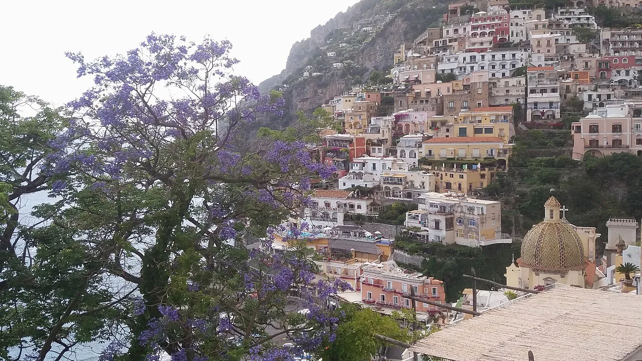 Purple jacaranda blossom framing Positano's hillside houses and the tiled dome of Santa Maria Assunta