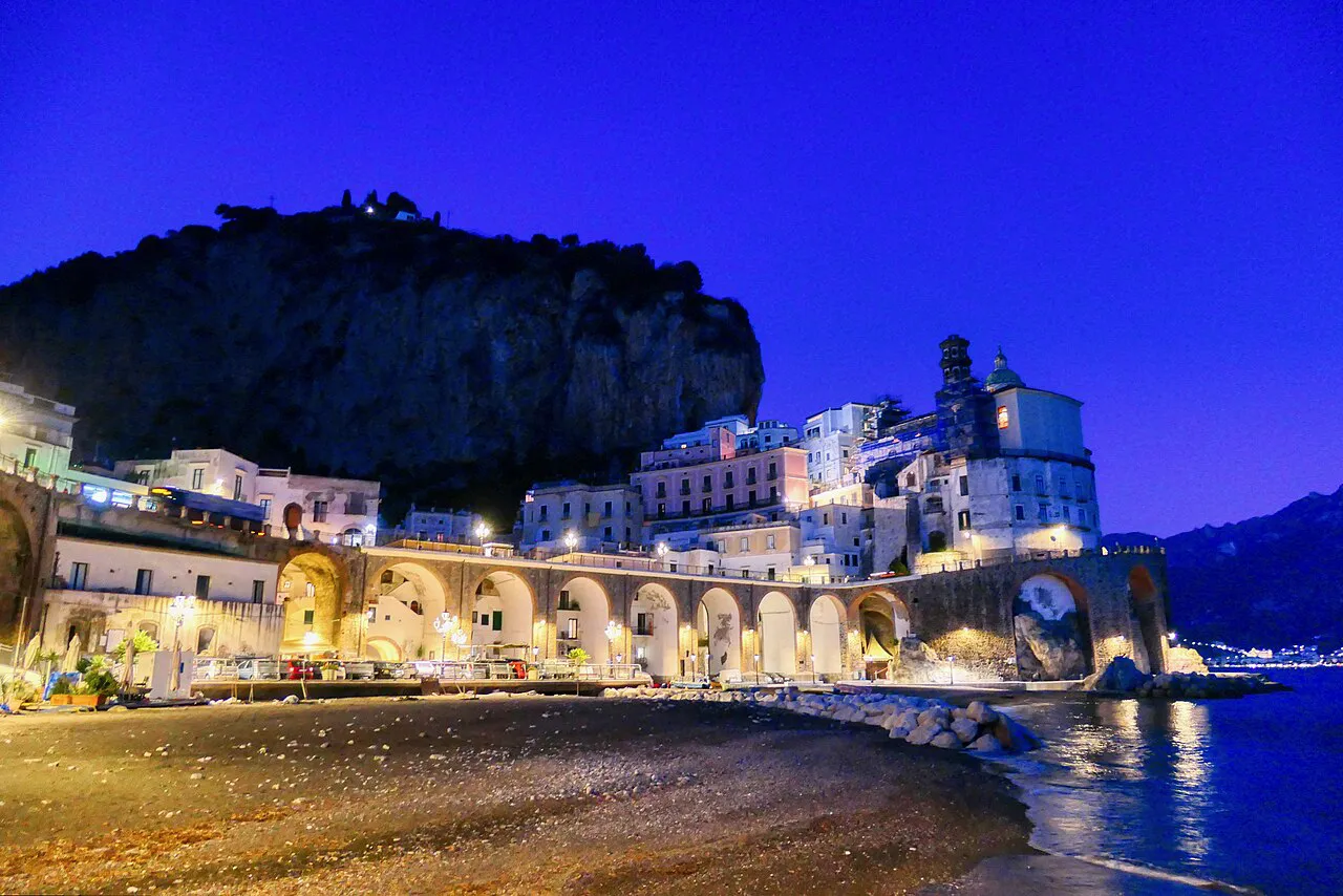 Atrani's small beach, arched seafront and church lit up at dusk beneath a dark cliff on the Amalfi Coast