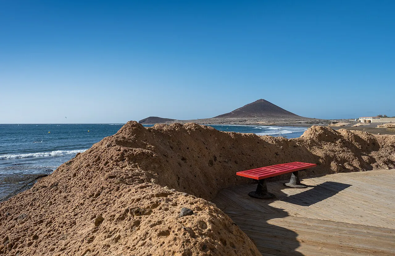 Boardwalk bench among sandstone rocks at El Médano beach with Montaña Roja on the horizon, Tenerife