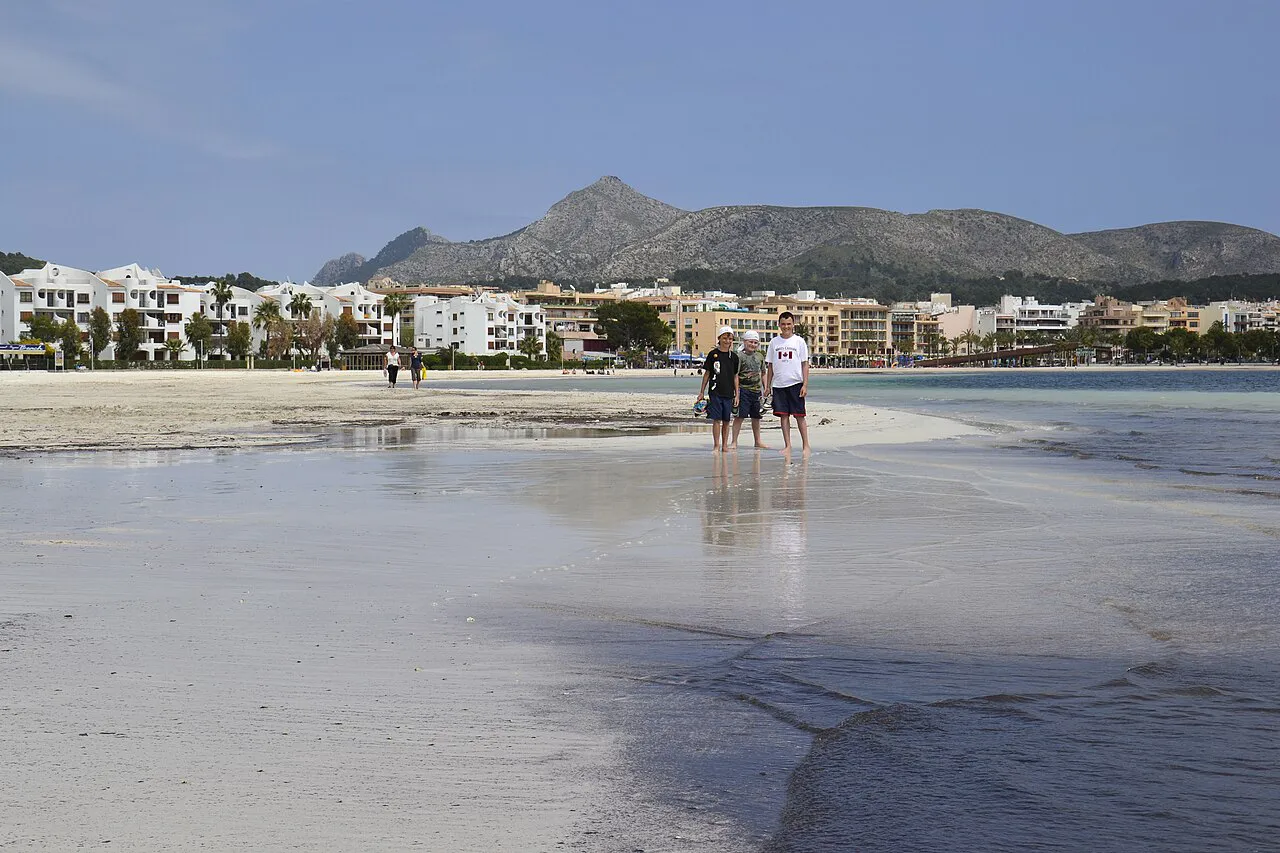 Wide shallow sands of Alcúdia beach reflecting the sky, with white seafront buildings and mountains beyond