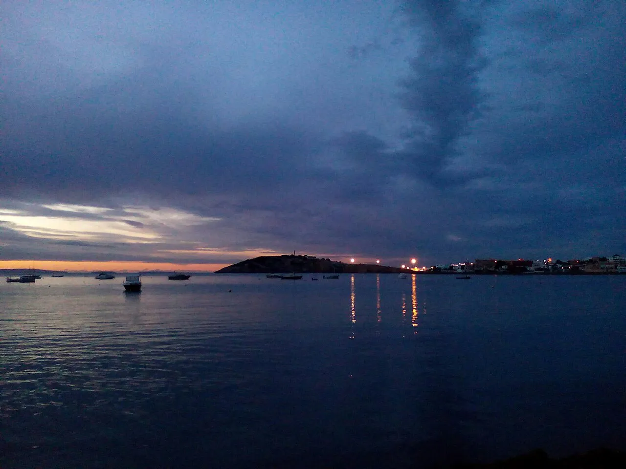 Storm clouds over Talamanca Bay at dusk, with moored boats and lights along the far shore of Ibiza