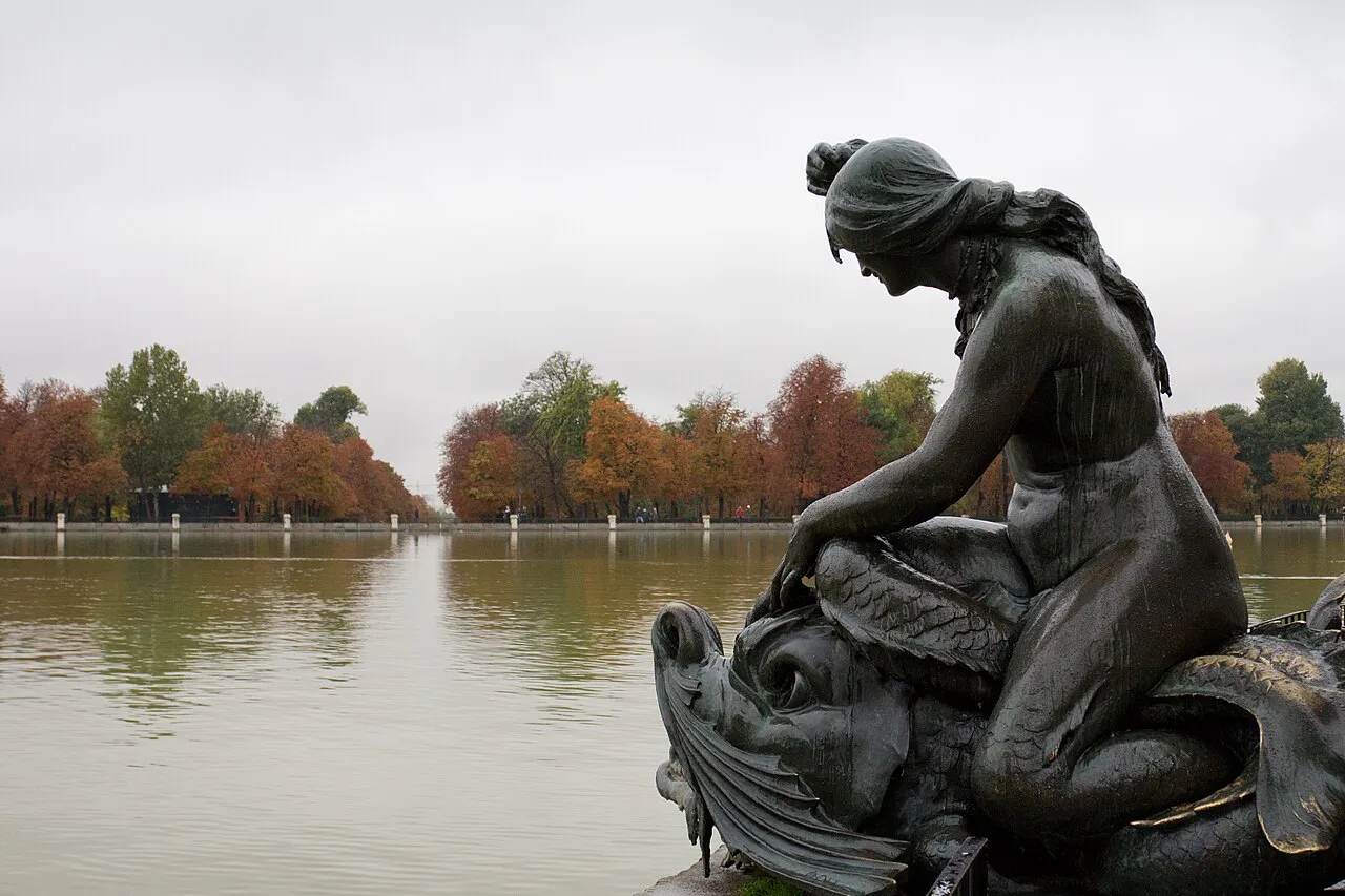 Bronze mermaid sculpture beside the Retiro Park pond in Madrid with autumn trees across the water