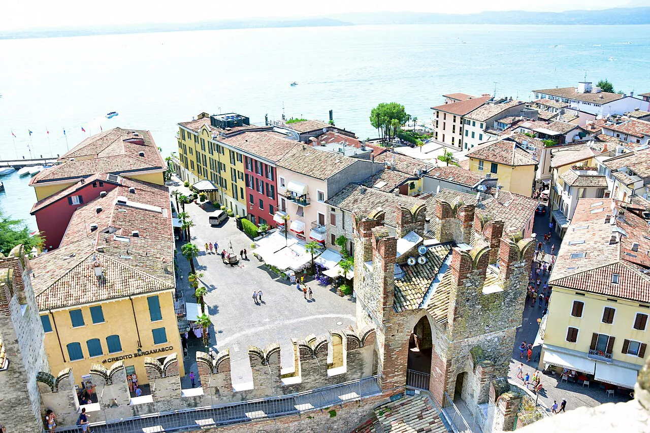 Colourful houses and a café-lined square inside Sirmione's old town, seen from above with Lake Garda behind