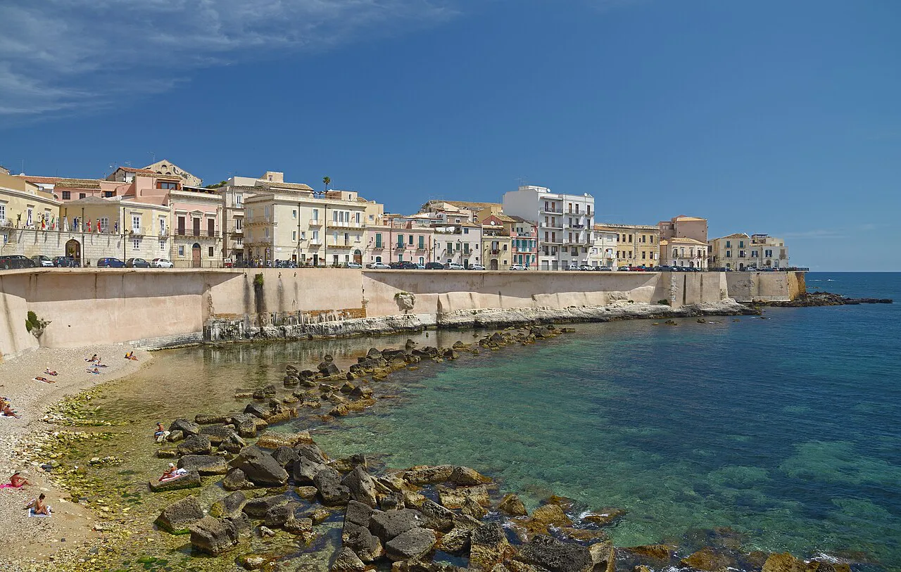 Pastel buildings above the sea wall and rocky shore of the Lungomare di Levante on Ortigia, Siracusa