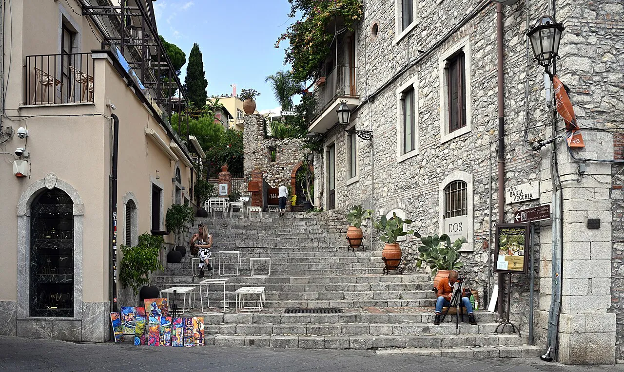 Stone steps of Salita Badia Vecchia climbing between old houses with potted plants in Taormina