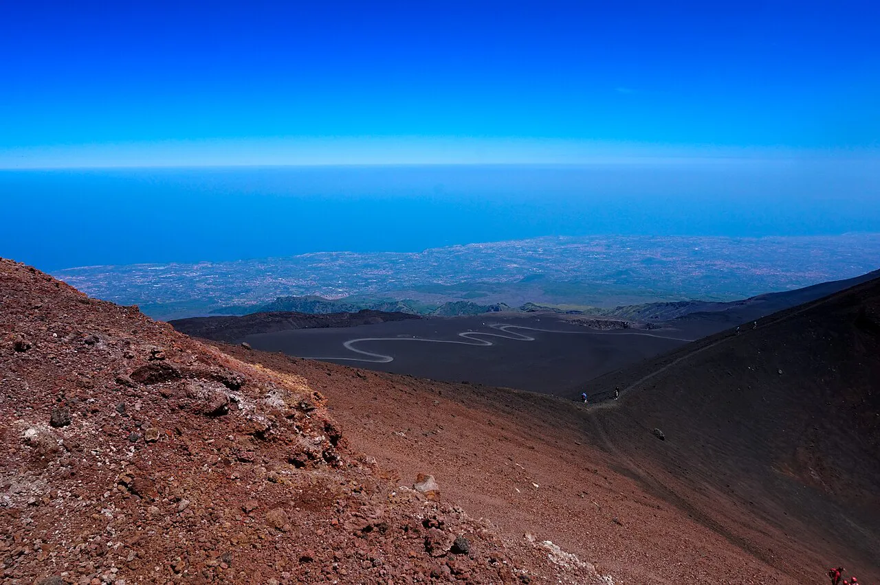 Red volcanic slopes on the summit of Mount Etna, with the plain and coast around Catania far below