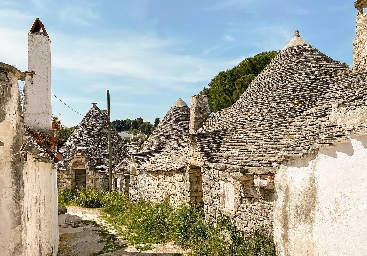 Weathered unpainted stone trulli with tall chimneys along a grassy path in Alberobello