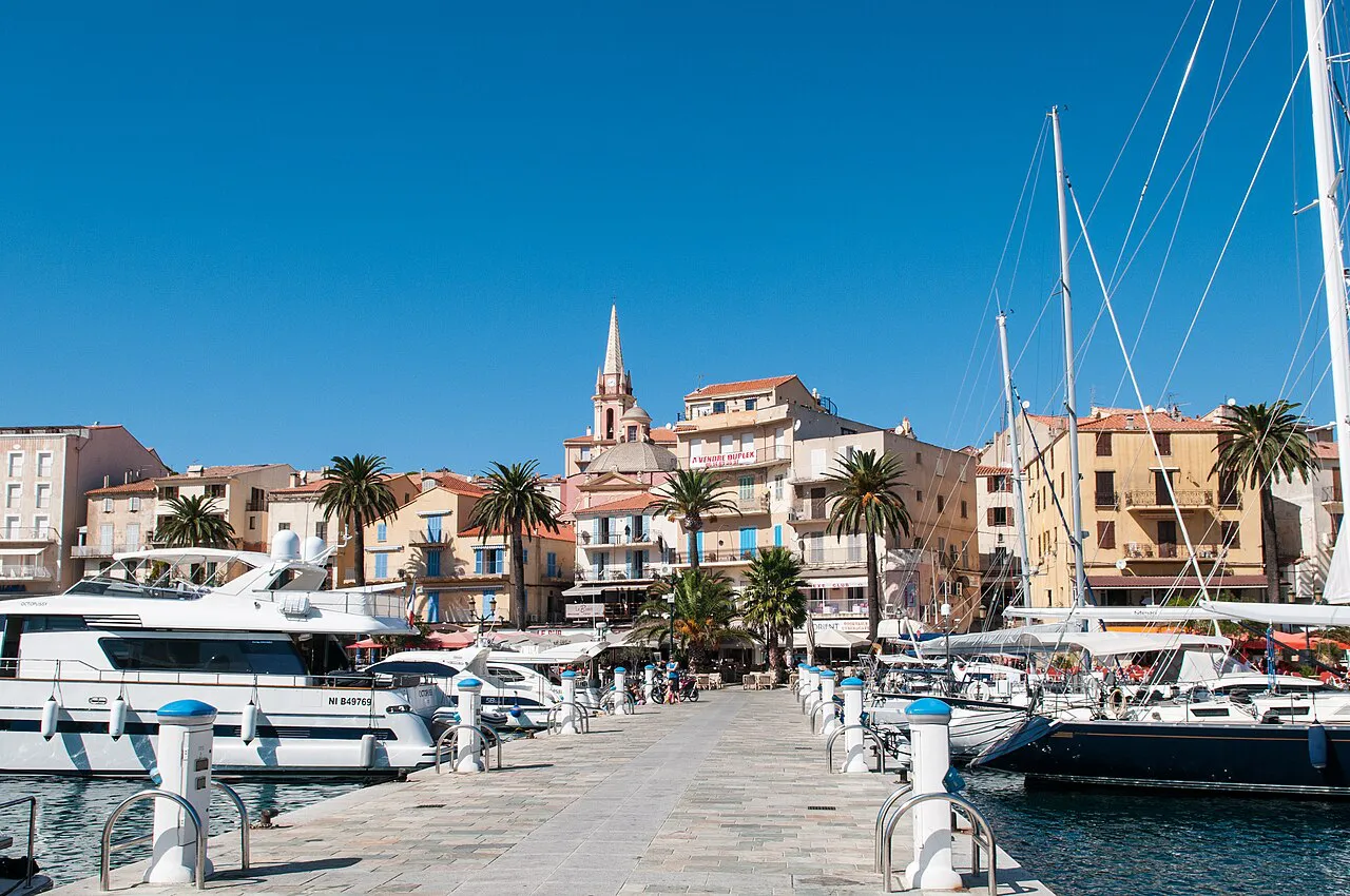 Yachts moored along the marina quay in Calvi with palm trees, pastel houses and a church spire