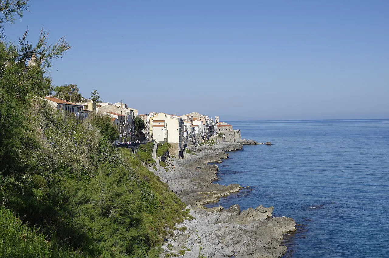 Rocky shoreline and whitewashed houses of Cefalù's old town along the calm blue sea