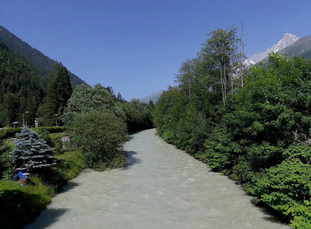 The glacial Arve river flowing between green trees near Les Bossons, with a snowy peak beyond