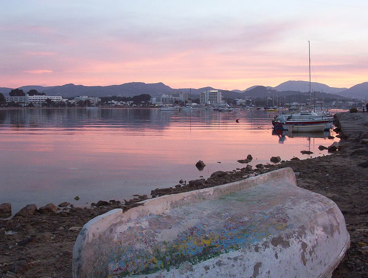 An old wooden boat on the rocky shore at Ibiza Town under a pink sunset sky