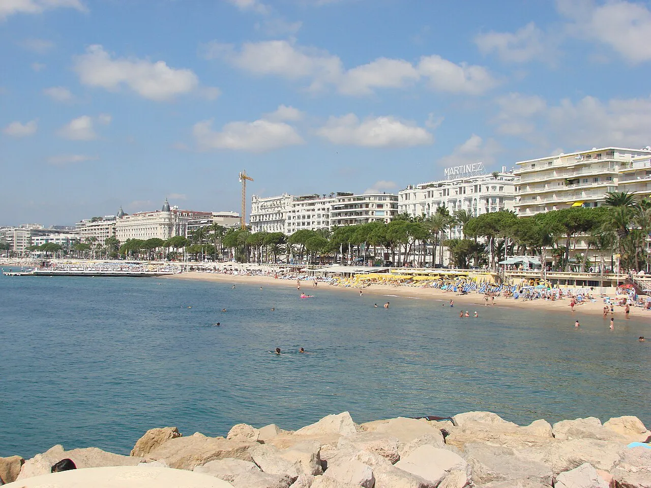 The Croisette seafront in Cannes seen across the bay from a rock breakwater, with beach and palms