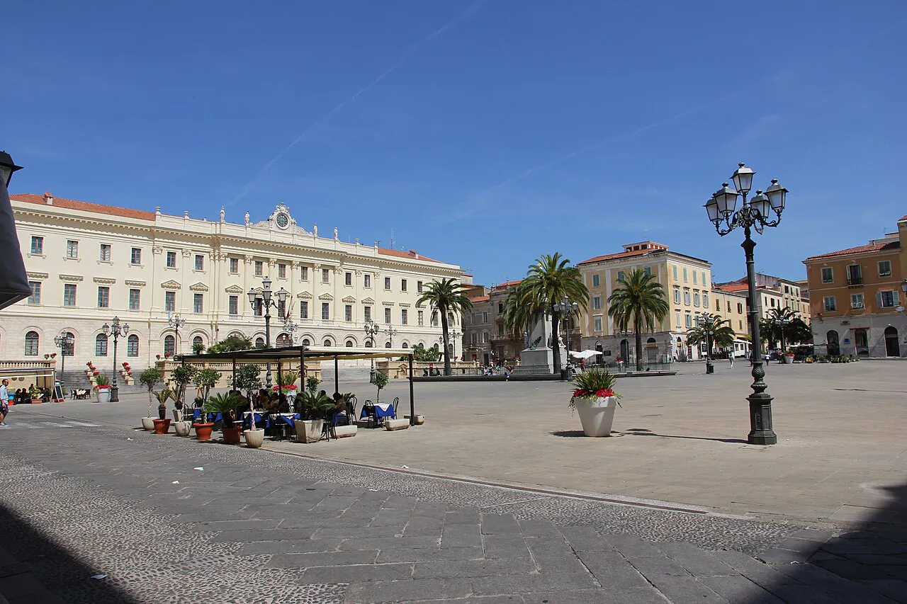 The broad paved Piazza d'Italia in Sassari with palm trees, ornate lamps and pale neoclassical palazzi