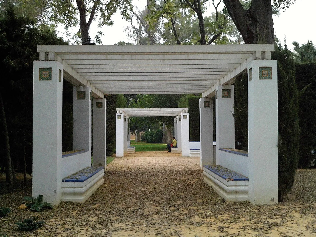 White pergola with tiled benches framing a gravel path in Parque de María Luisa, Seville