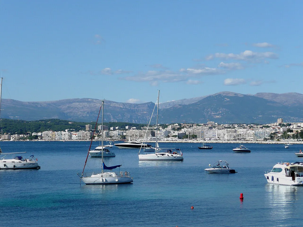 Sailing boats anchored in the calm bay, with the Juan-les-Pins seafront and hills behind, seen from Cap d'Antibes