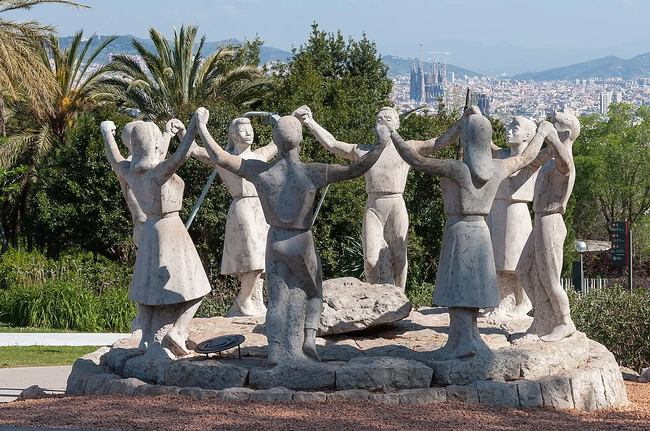 Stone Sardana dancers sculpture in a Montjuïc garden with palms and the Barcelona skyline beyond
