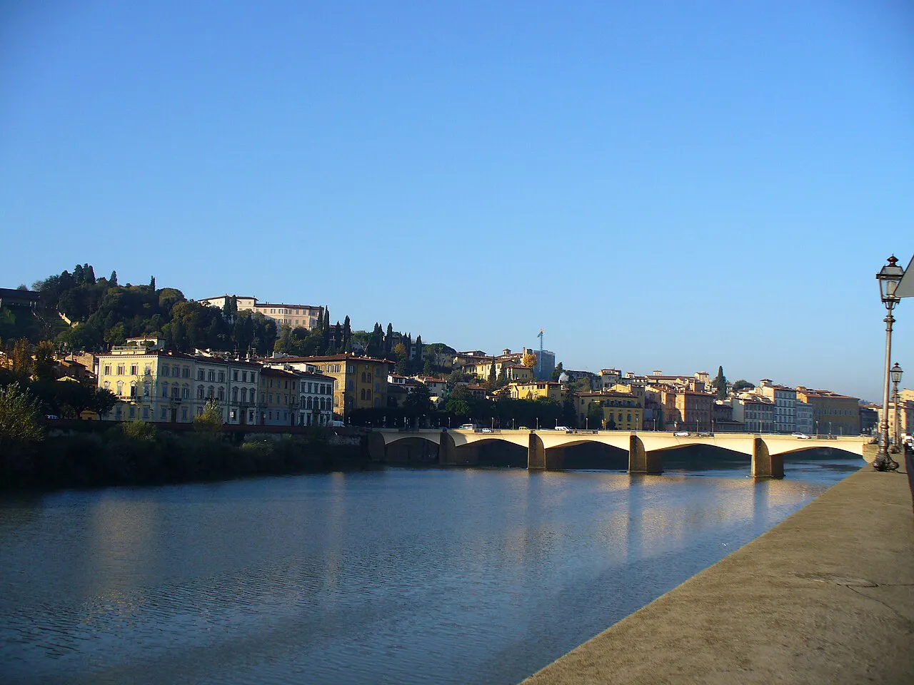 The calm Arno in Florence under a blue sky, with a stone bridge and hillside villas beyond