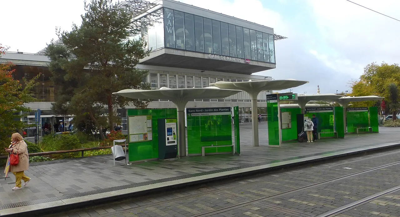 Tram stop with green shelters on the forecourt of Nantes railway station, the glass station building behind