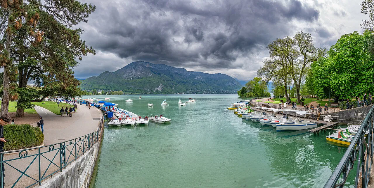 Tree-lined lakeside promenade and moored boats on the turquoise water of Lake Annecy under stormy clouds