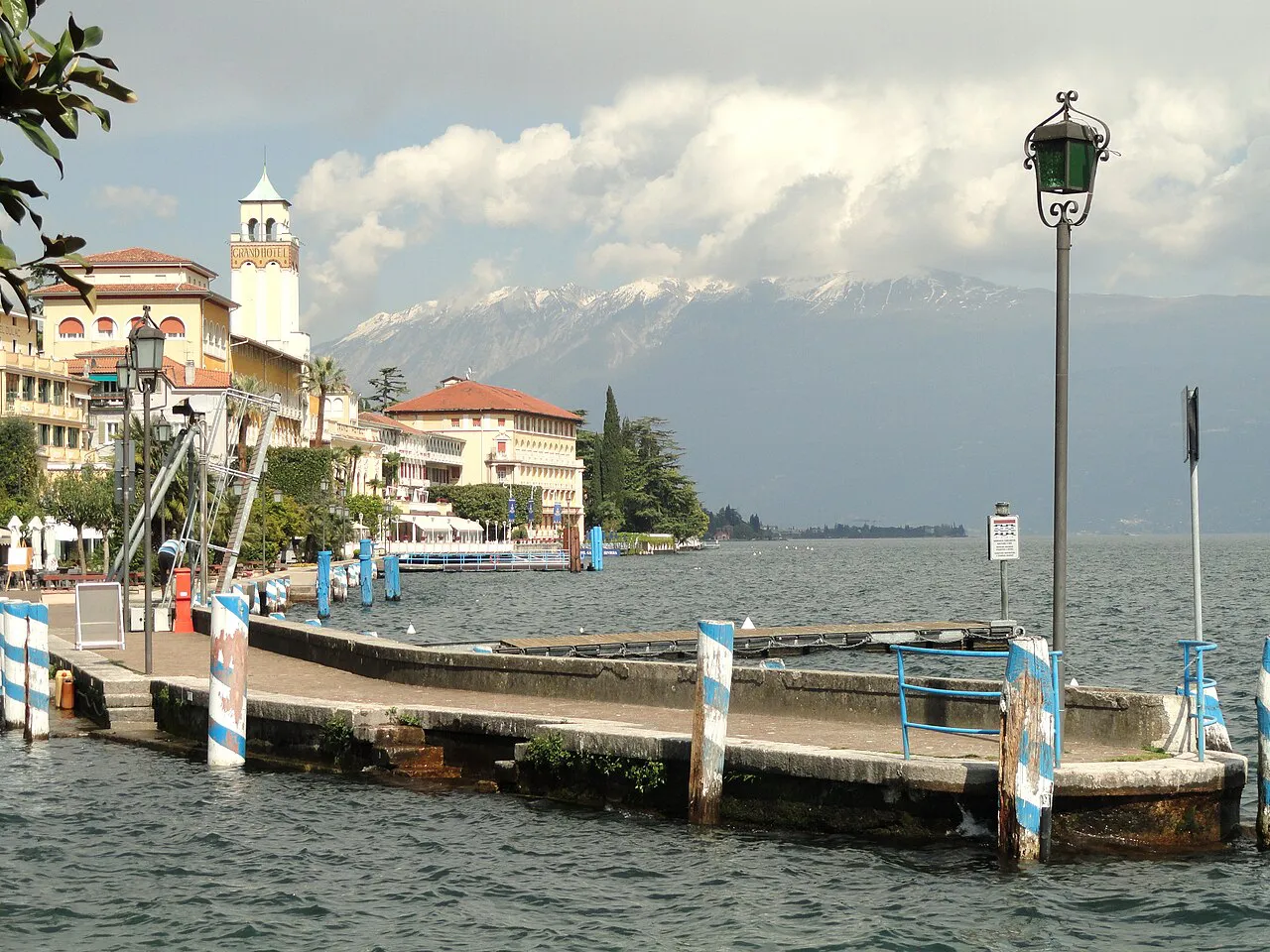 Boat pier and lakeside promenade at Gardone Riviera with snow-capped mountains across Lake Garda