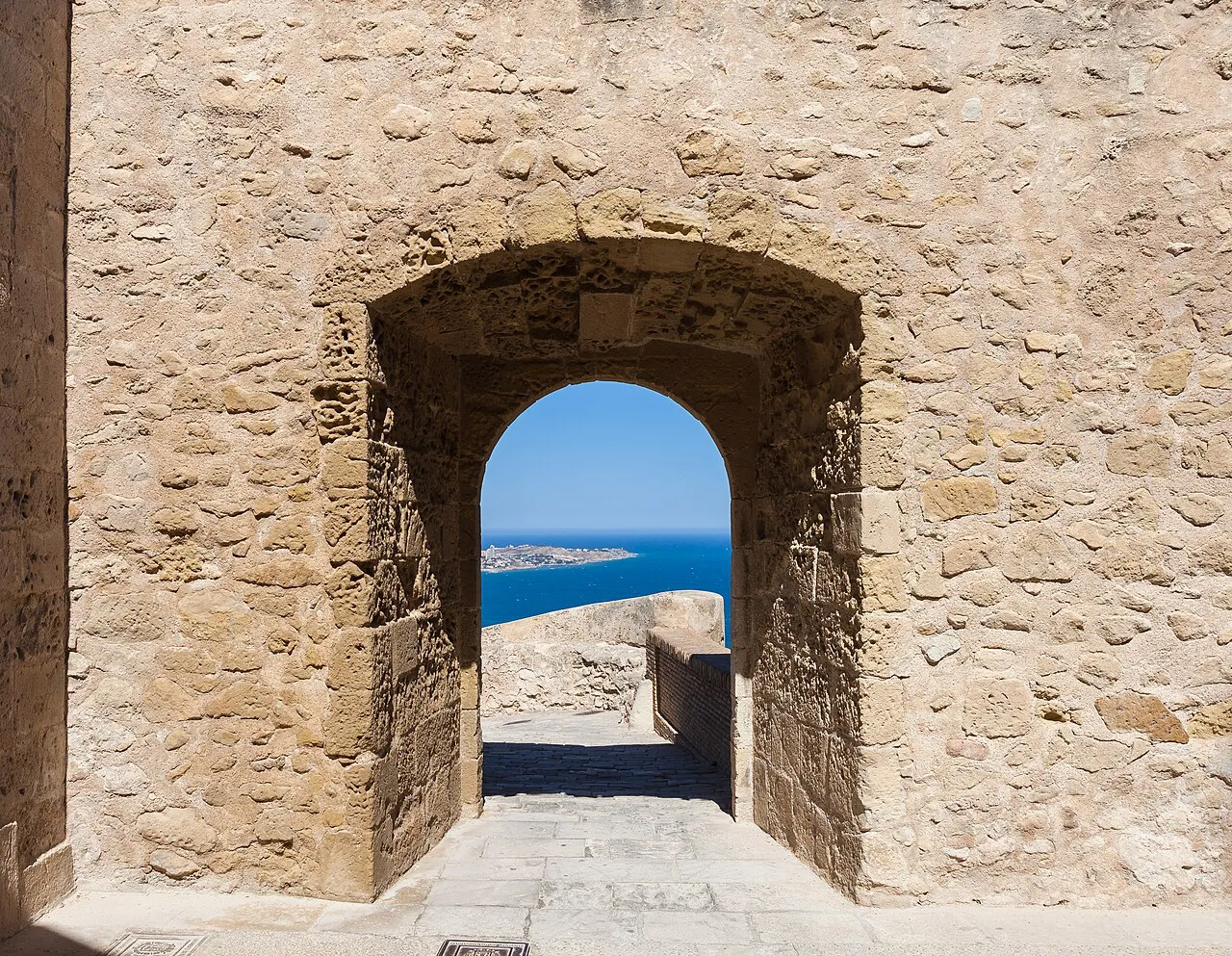 Stone archway in the walls of Santa Bárbara Castle in Alicante framing a glimpse of the blue sea