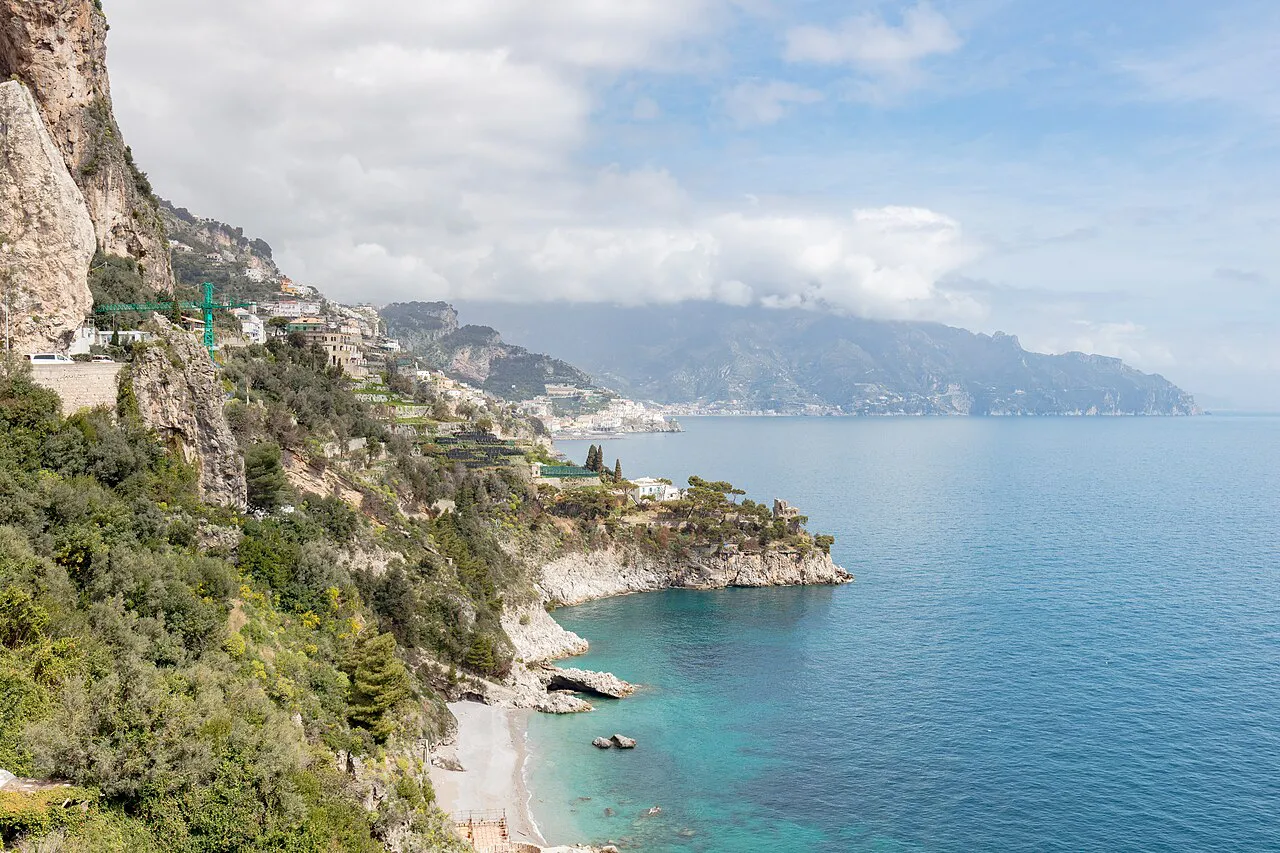 Green cliffs and a small turquoise cove along the coast at Conca dei Marini, with the shoreline beyond
