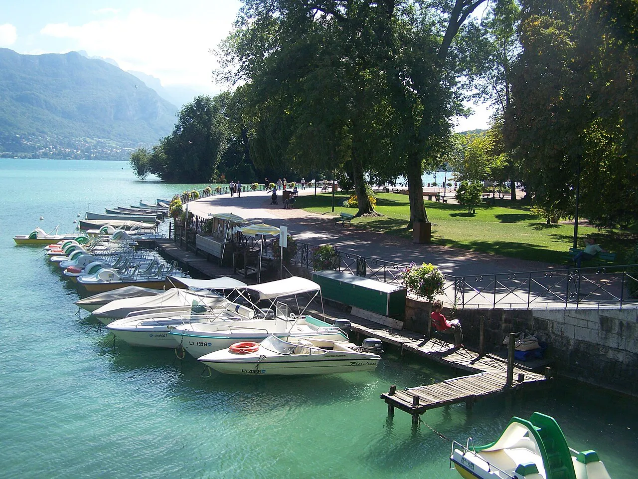Moored boats along the shore of Lake Annecy beside the Jardins de l'Europe, with mountains across the water