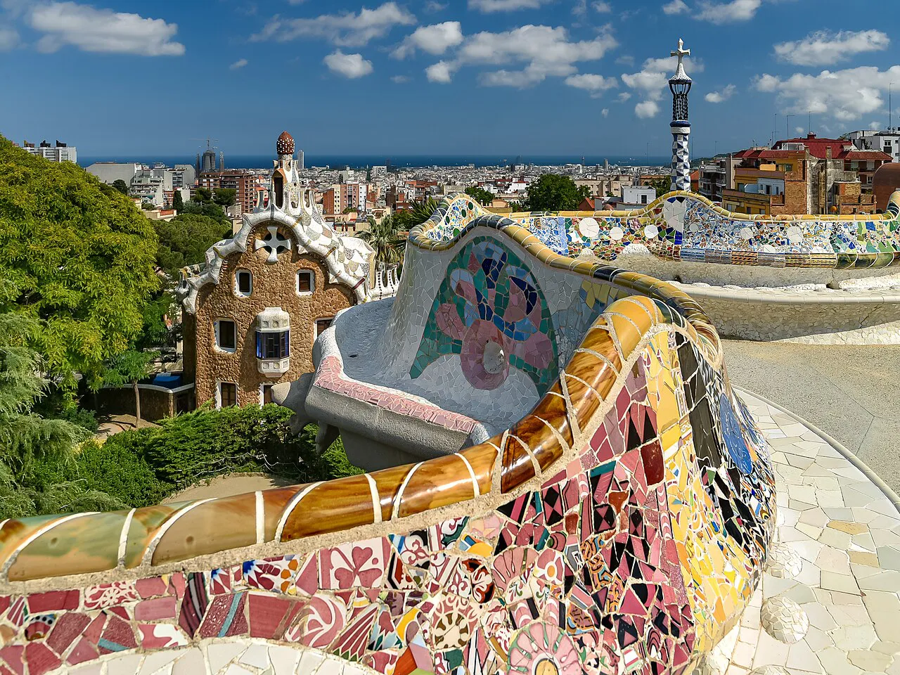 Mosaic-tiled serpentine bench and gatehouse spire at Park Güell with Barcelona and the sea beyond