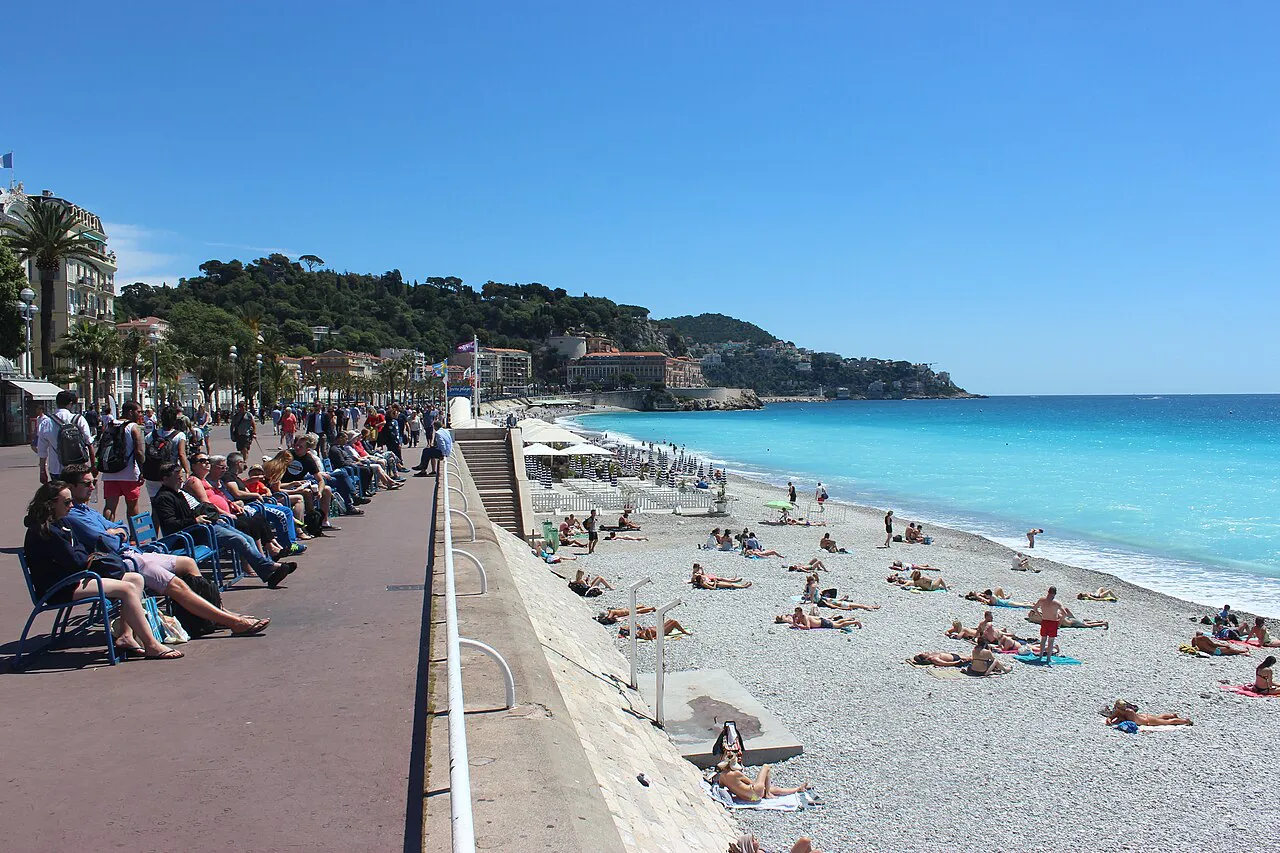 Promenade des Anglais in Nice beside a pebble beach and turquoise sea, with Castle Hill in the distance