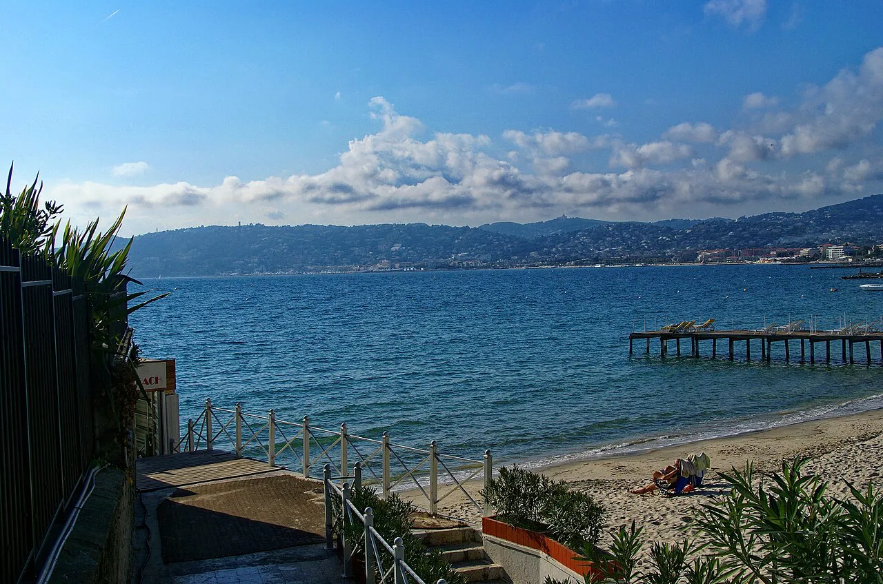Sandy beach and wooden jetty at Juan-les-Pins on a bright day, with Cap d’Antibes across the bay