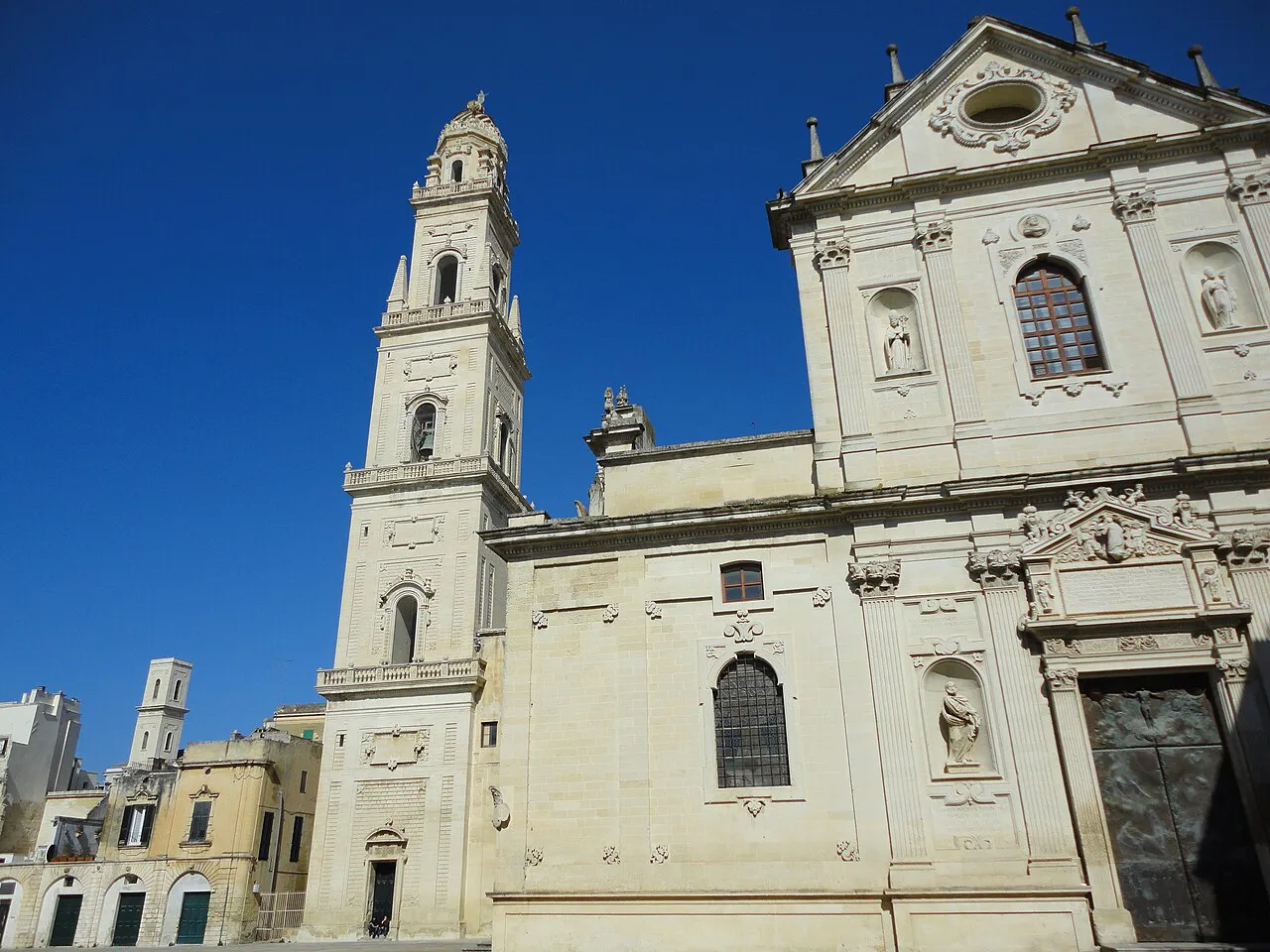 White Baroque facade of Lecce Cathedral beside its tall bell tower against a deep blue sky