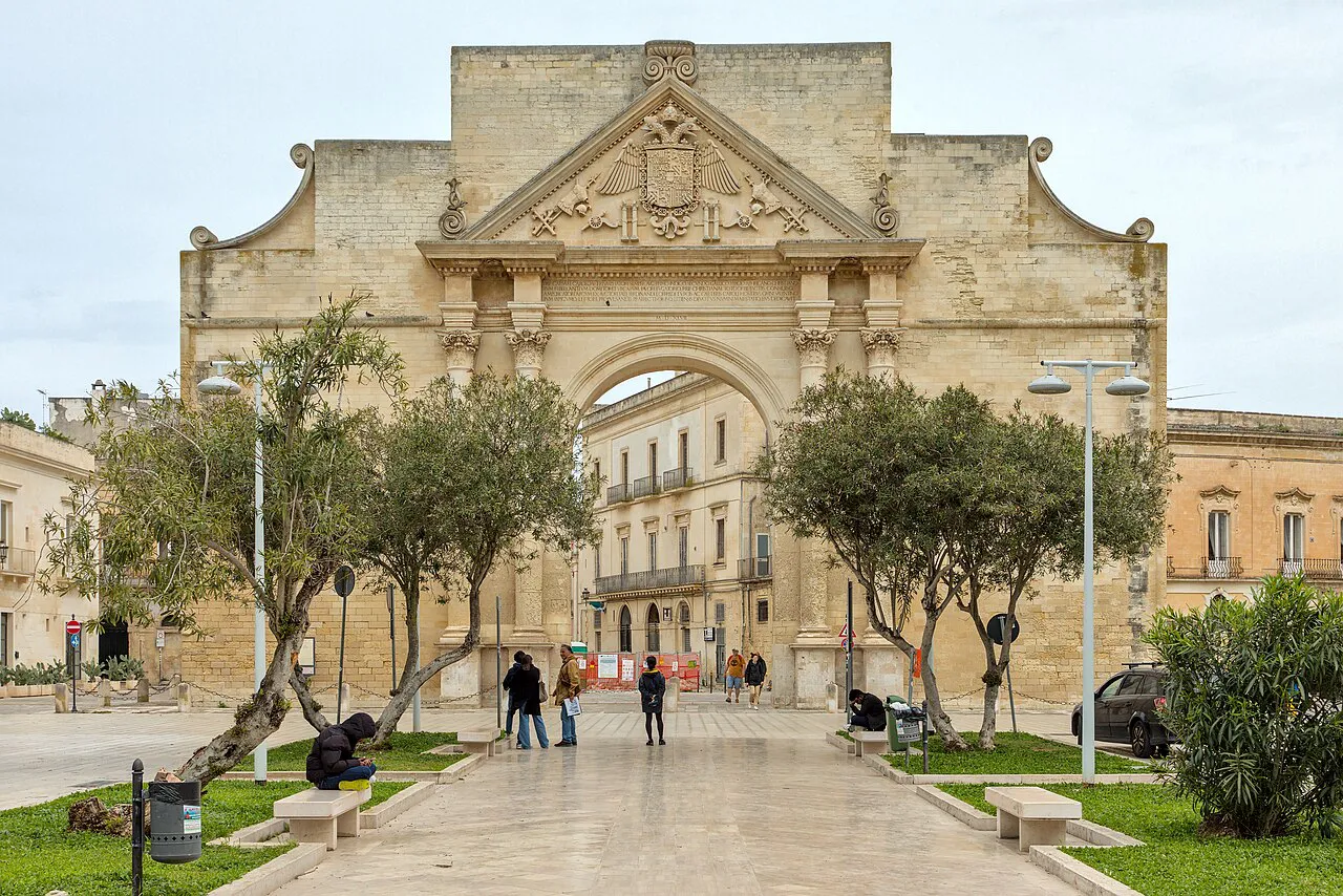 Porta Napoli triumphal arch in Lecce seen along a paved avenue lined with olive trees
