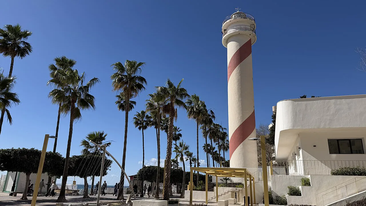 Red-and-white striped Marbella lighthouse among tall palm trees on the seafront promenade