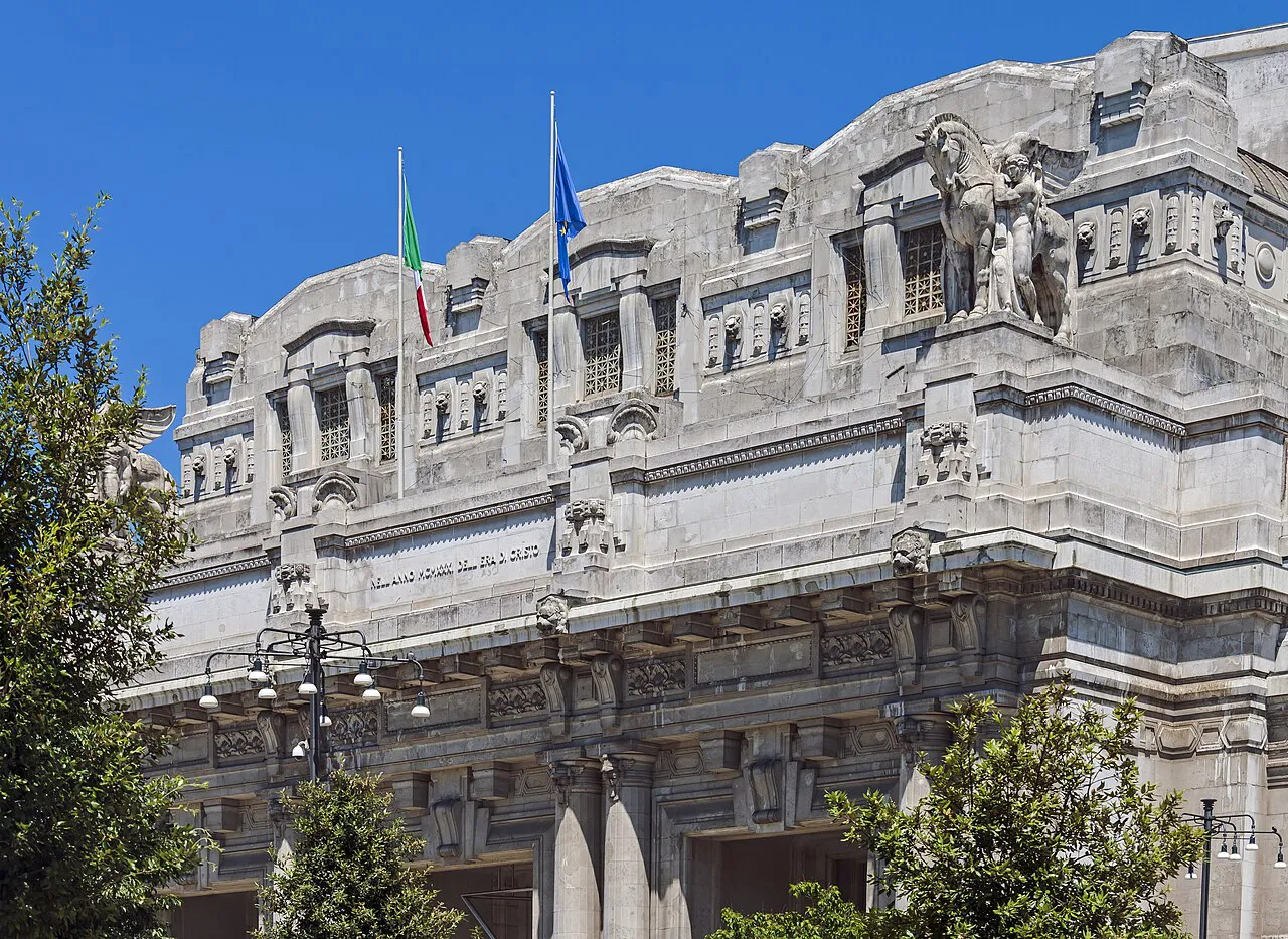 Carved upper facade of Milano Centrale railway station with flags against a blue sky