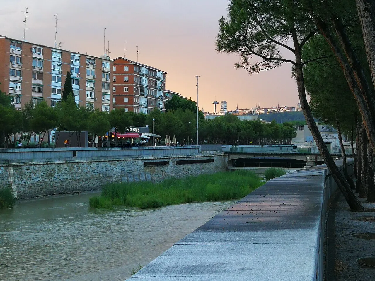 Riverside walkway along the Manzanares in Madrid Río park at sunset, with pines and apartment blocks