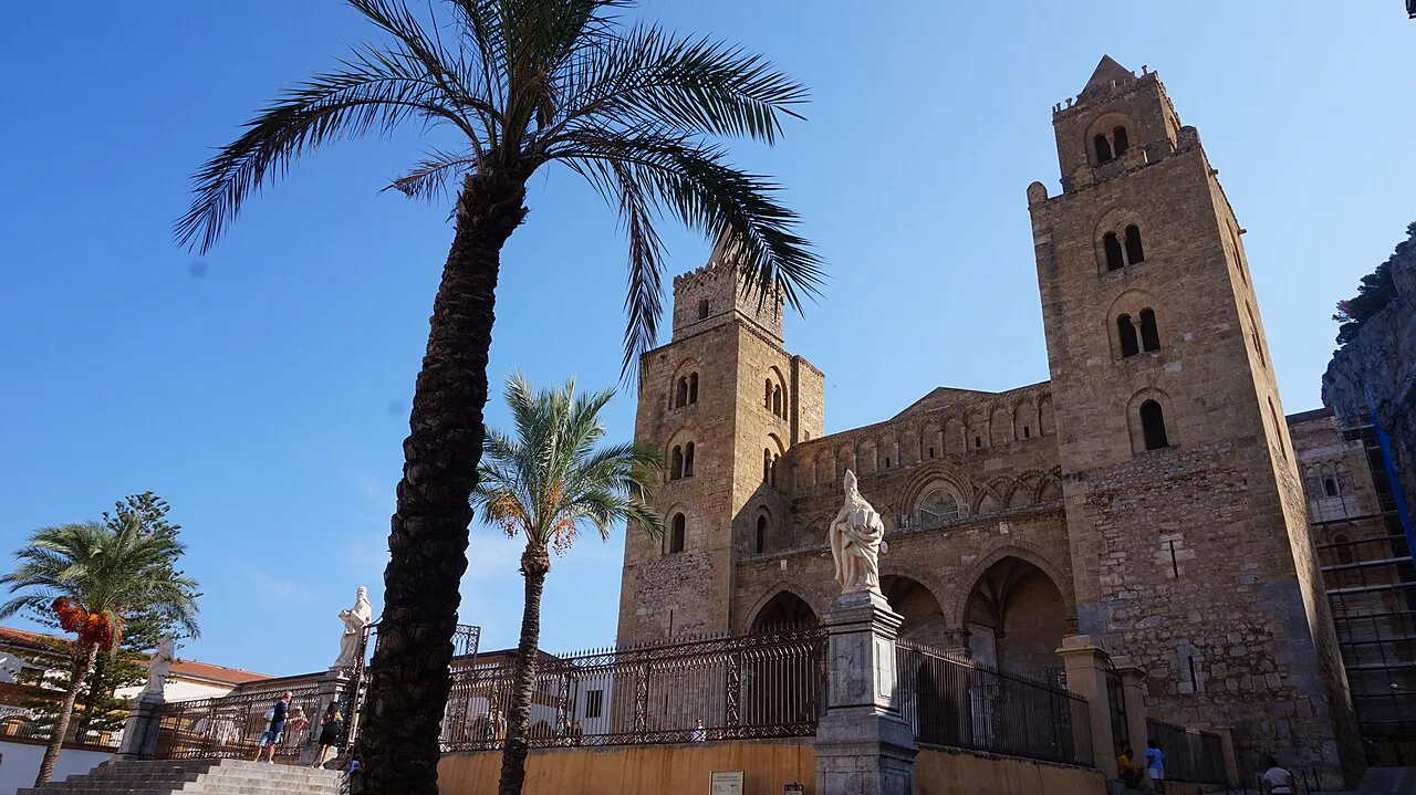 The twin Norman towers and facade of Cefalù Cathedral framed by tall palm trees under a blue sky, Sicily