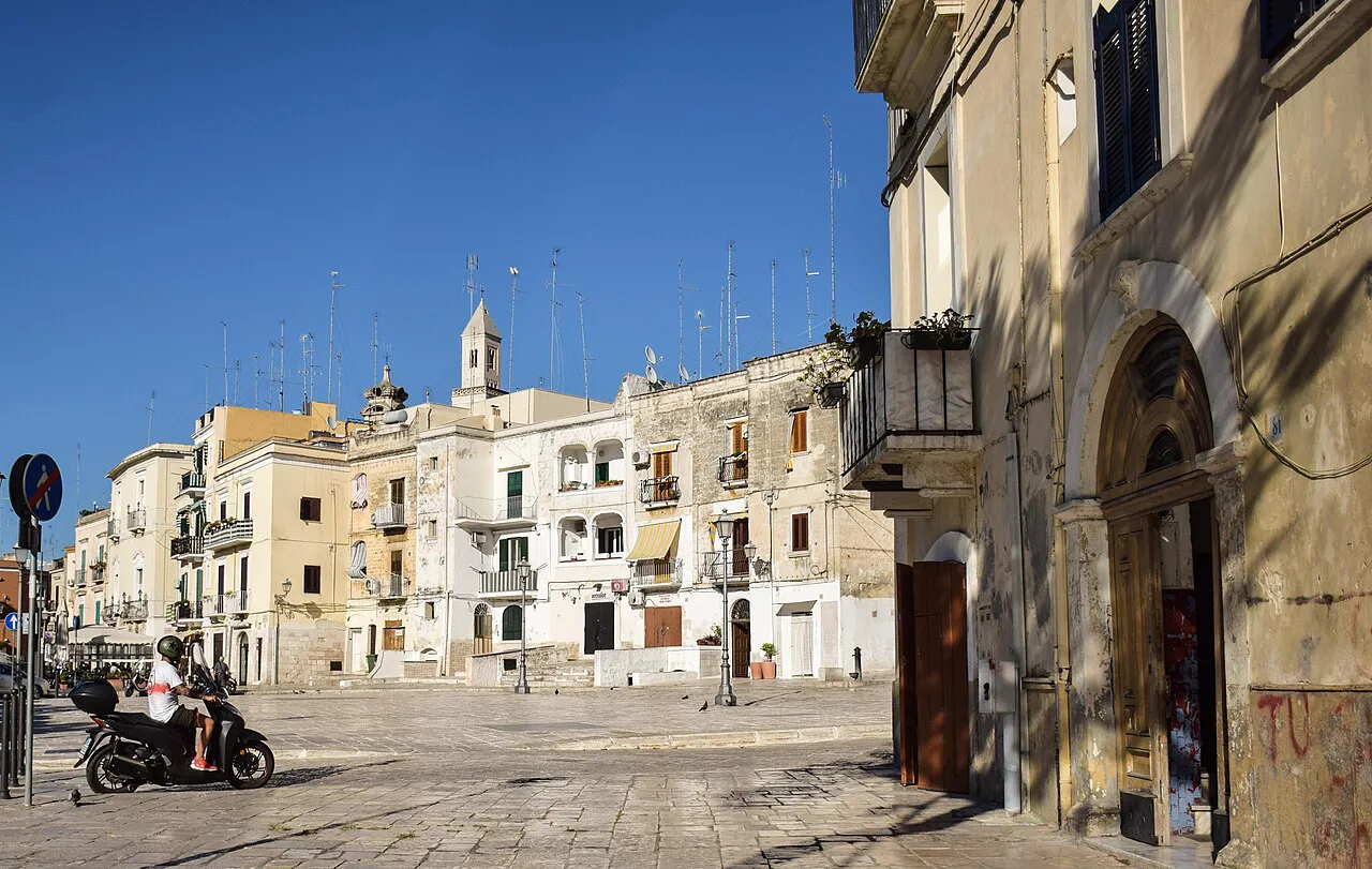 Pale stone houses and a church bell tower around a sunny paved square in Bari Vecchia