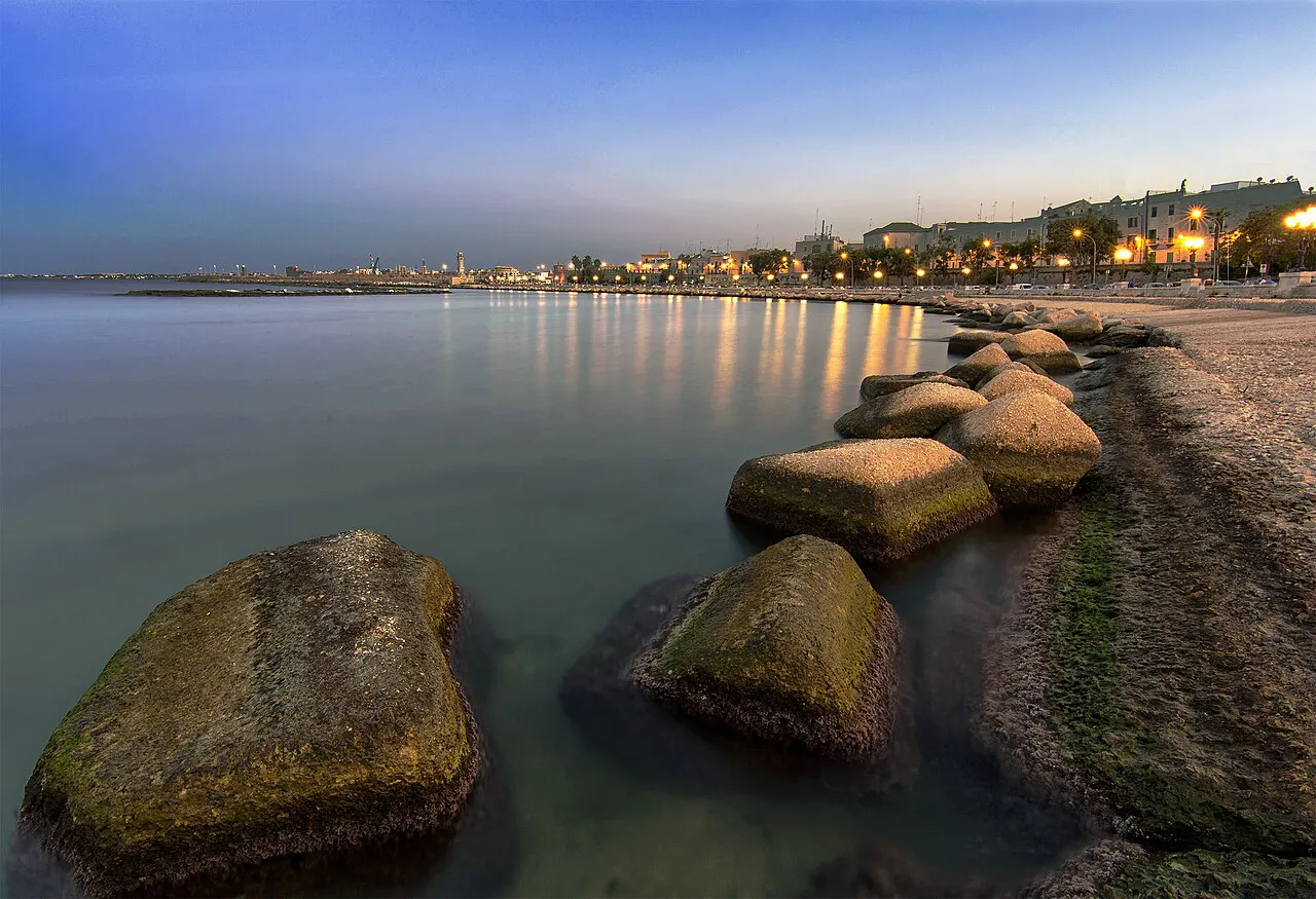 Smooth boulders in calm water along the Bari seafront at dusk, with city lights curving along the shore