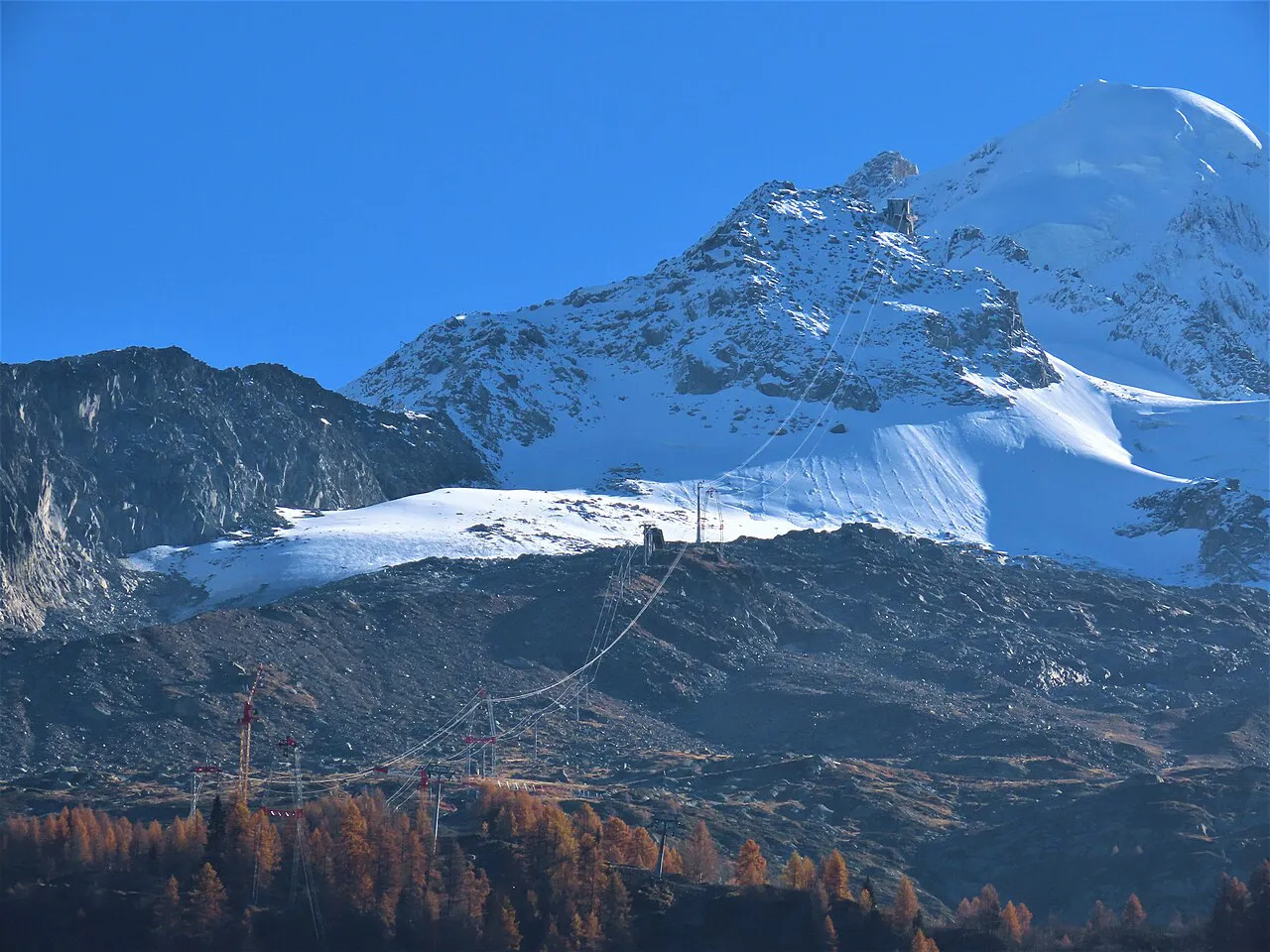 Cable car pylons climbing toward the snow-covered Aiguille des Grands Montets above autumn larches at Argentière