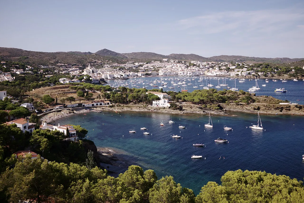 Sheltered cove with moored sailboats below pine trees, with the white town of Cadaqués across the bay