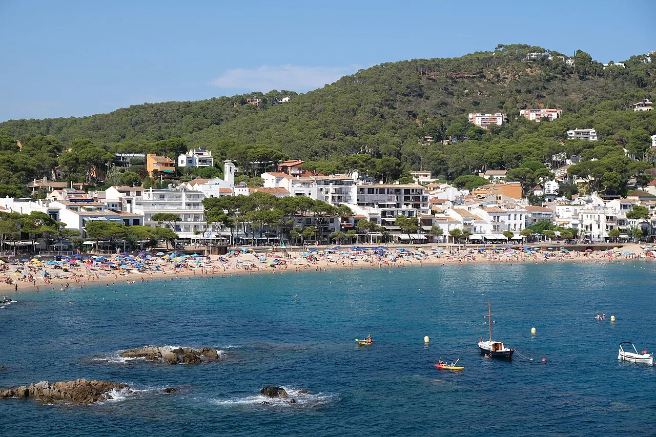 Llafranc's curved sandy beach and white village below wooded hills, seen across the bay
