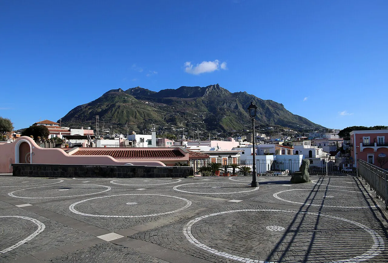 Paved piazza in Forio with white houses and the green slopes of Monte Epomeo rising behind, Ischia