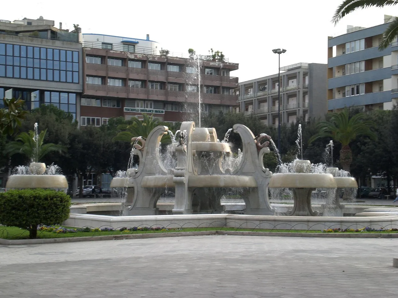 Sculpted fountain in Piazza Mazzini, Lecce, with palms and modern buildings around the square