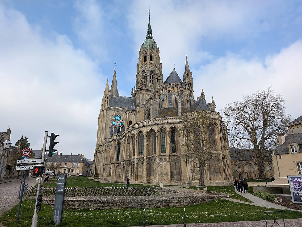 Bayeux Cathedral’s Gothic chevet and central tower seen across a lawn under a cloudy blue sky