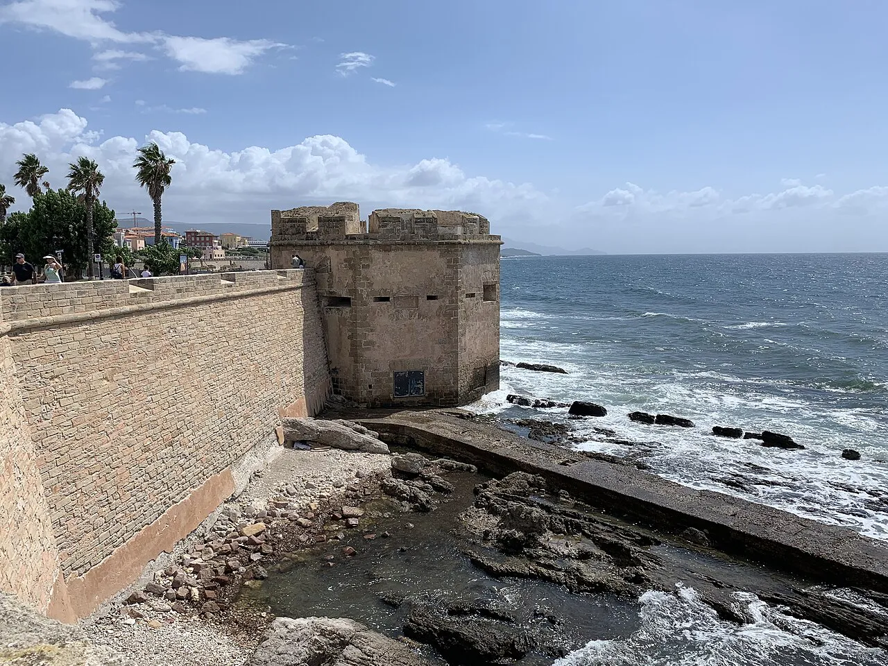 A stone tower on Alghero's sea walls above rocks and breaking waves on a bright day