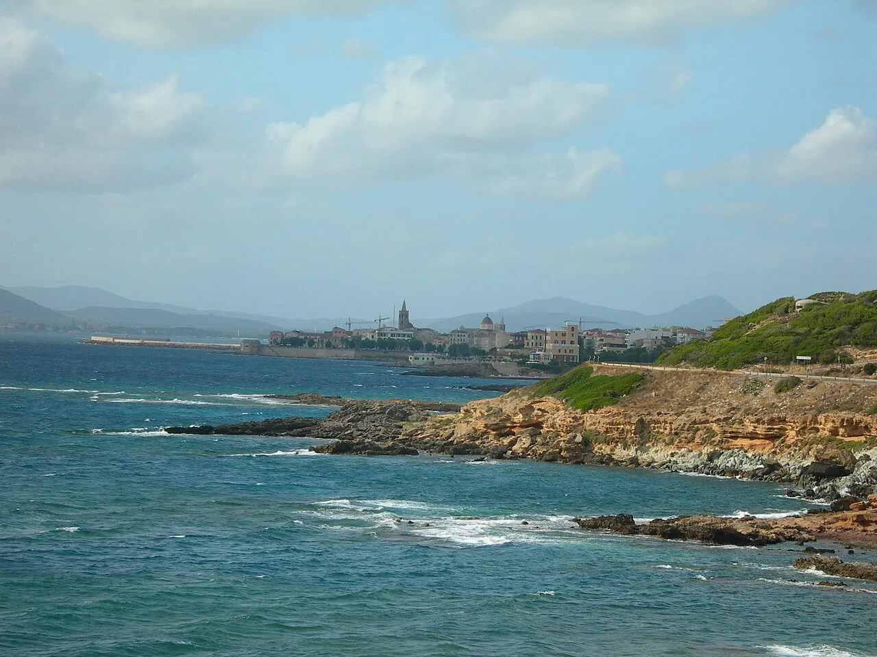 Alghero's old town on the waterfront seen across rocky coves and turquoise sea